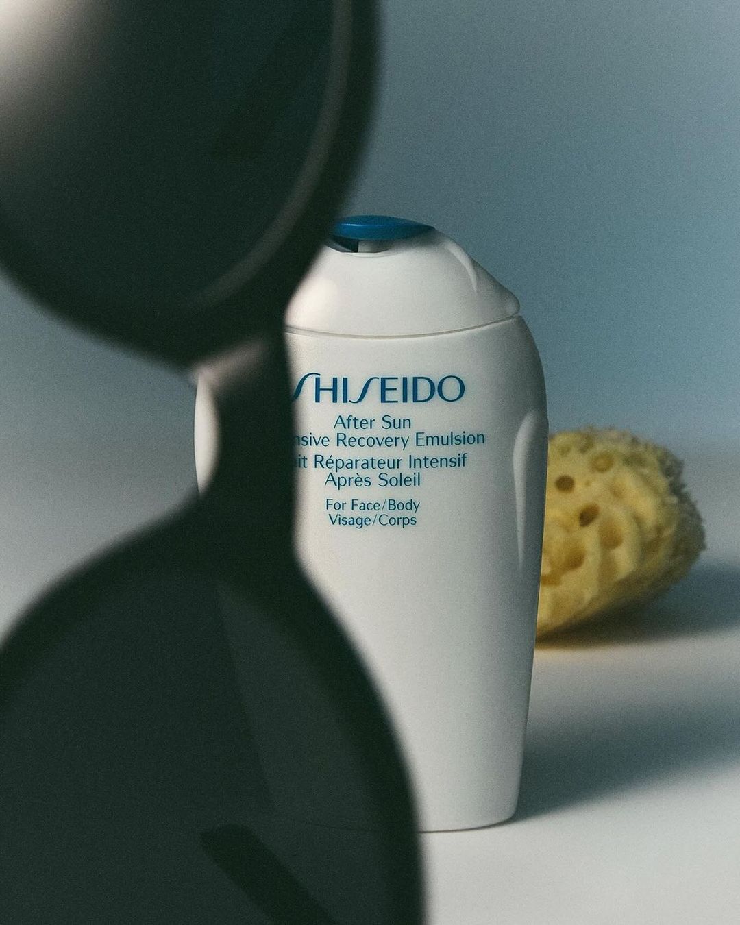 Shiseido After Sun Intensive Recovery Emulsion bottle is showcased alongside a sea sponge, partially obscured by a dark hourglass.