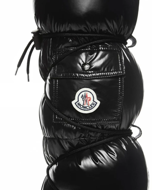 Close-up of black puffy snow boots with black laces and Moncler logo, set against a white background, shiny, puffy texture.