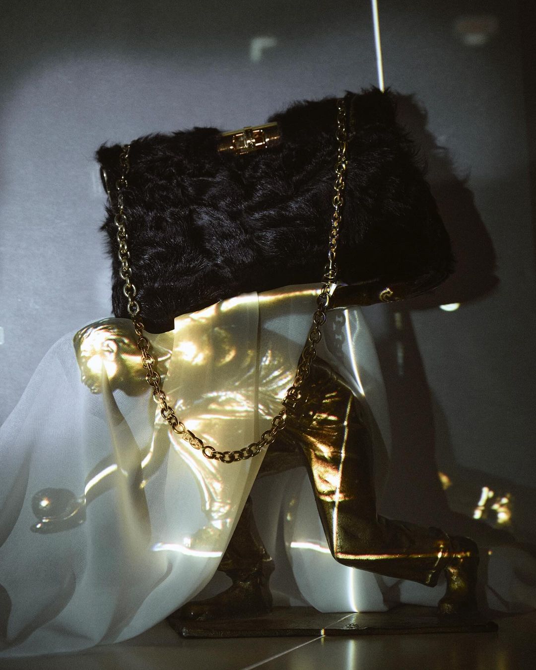 Black furry handbag with gold chain on a gold statue of person kneeling with white fabric backdrop in a studio setting.