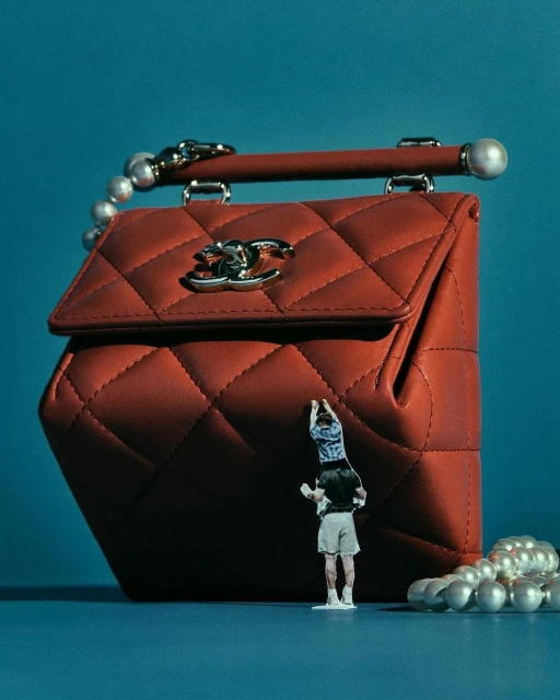 Red Chanel quilted handbag with pearl handle and miniature human figure, set against a blue backdrop in a studio setting.