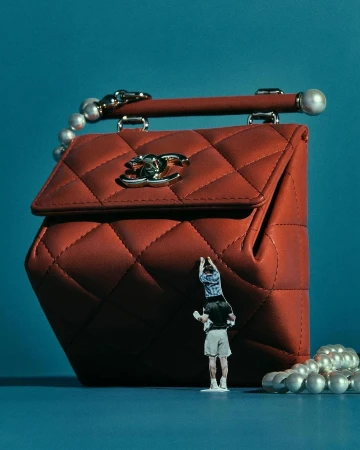 Red Chanel quilted handbag with pearl handle and miniature human figure, set against a blue backdrop in a studio setting.