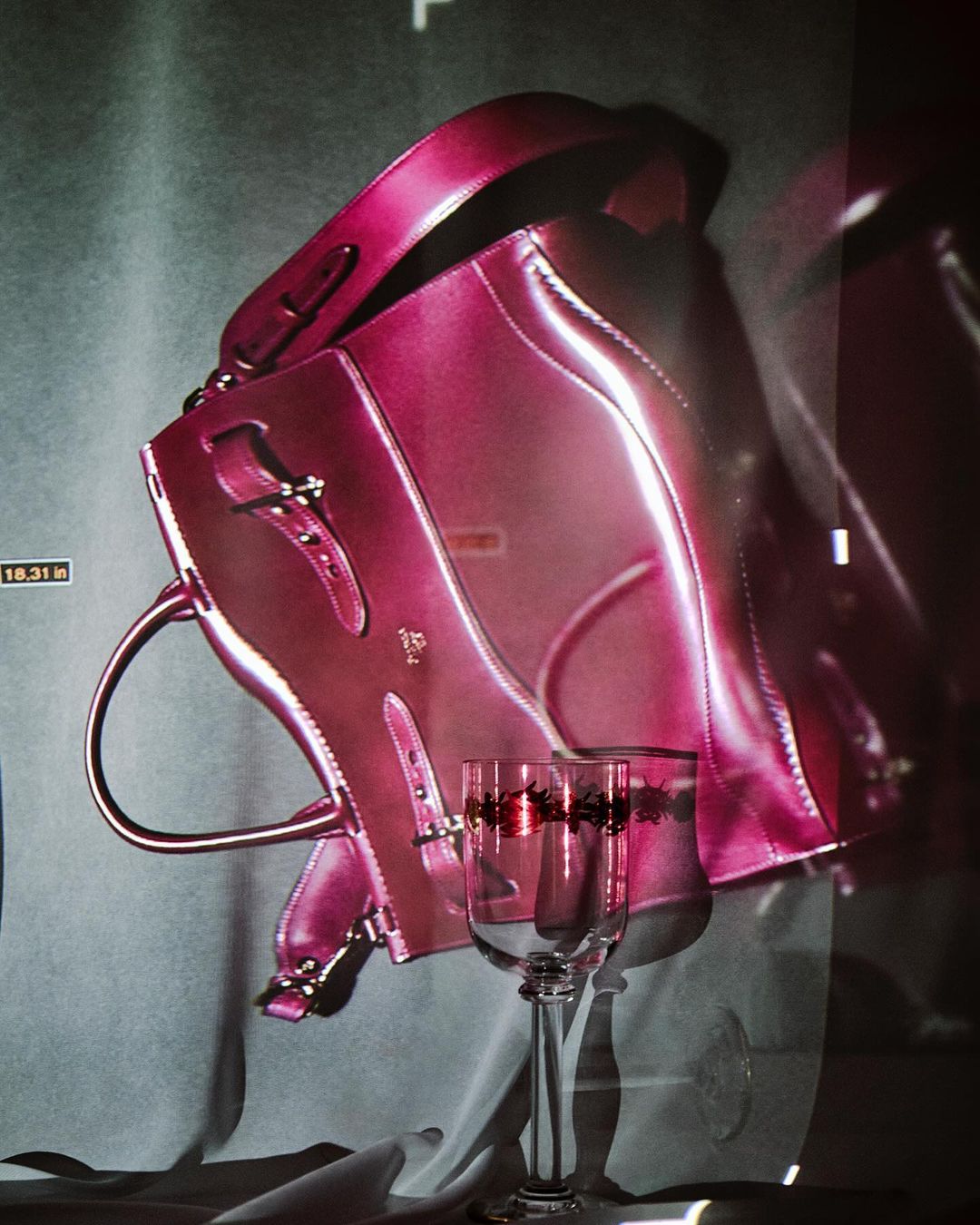 Still life image of a shiny, magenta-colored satchel-style bag with metal hardware, set against a mottled gray backdrop with a wine glass.