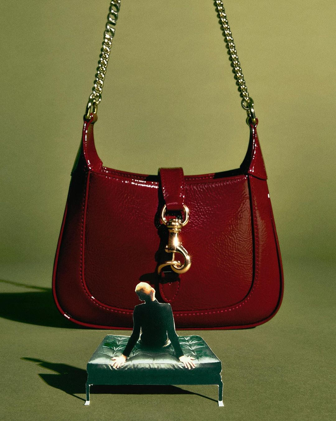 Burgundy patent leather purse with chain strap, small green tufted ottoman, and miniature man for an artful fashion statement.