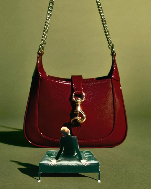 Burgundy patent leather purse with chain strap, small green tufted ottoman, and miniature man for an artful fashion statement.