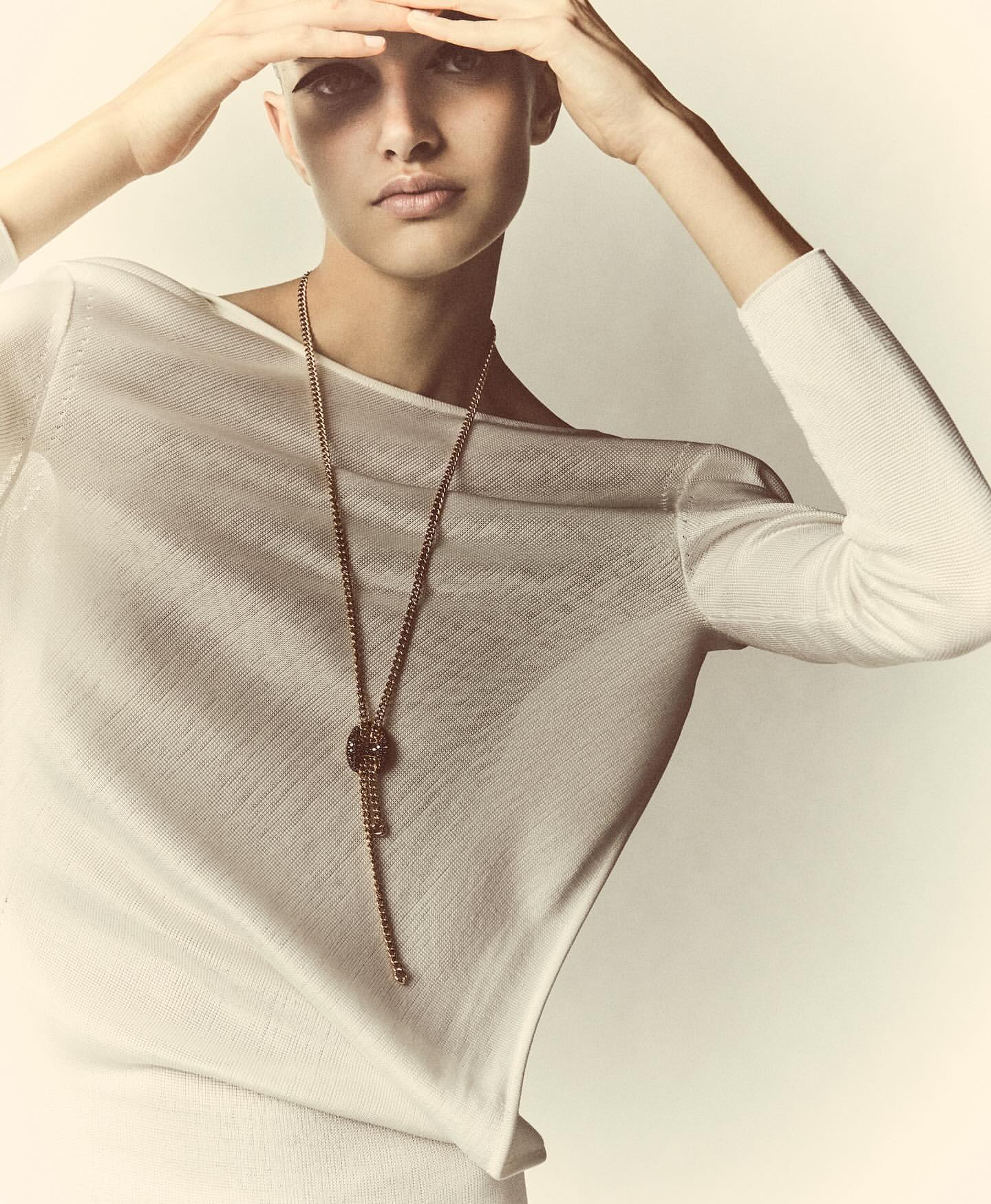 Woman in white top poses with hands above head, wearing a long beaded necklace against a soft white background in a fashion style.