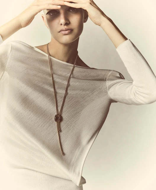 Woman in white top poses with hands above head, wearing a long beaded necklace against a soft white background in a fashion style.