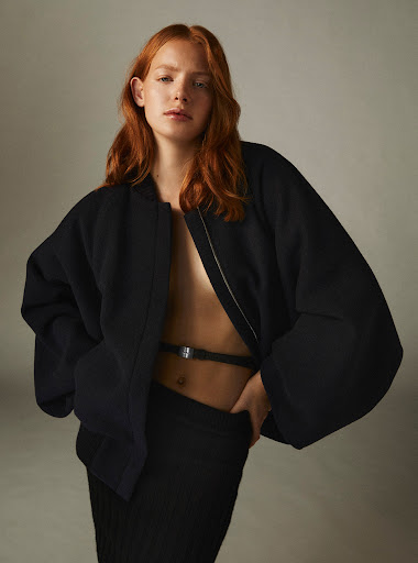 Red-haired female model wearing an oversized black bomber jacket and dark skirt against a neutral studio backdrop.