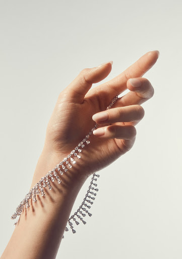 Woman's hand delicately holding a sparkling diamond necklace against a pale backdrop, accentuating the jewelry's radiant beauty and design.