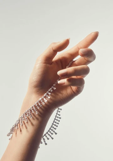 Woman's hand delicately holding a sparkling diamond necklace against a pale backdrop, accentuating the jewelry's radiant beauty and design.