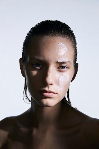 Close-up portrait of a woman with slicked-back hair and skincare product on her face against a bright white background.