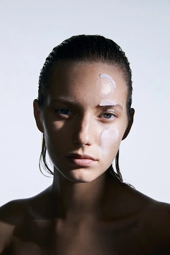 Close-up portrait of a woman with slicked-back hair and skincare product on her face against a bright white background.