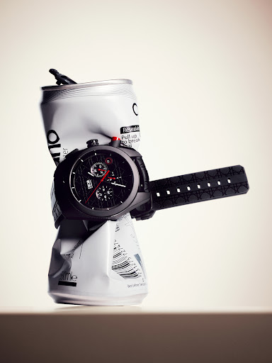 A black chronograph watch wrapped around a crushed white aluminum can against a plain gradient studio background.