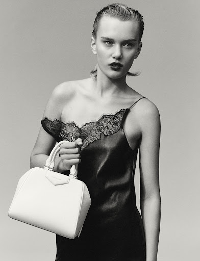 Studio shot of a young woman in a black lace-trimmed slip dress, holding a white satchel bag, with wet-look hair and dark lipstick.