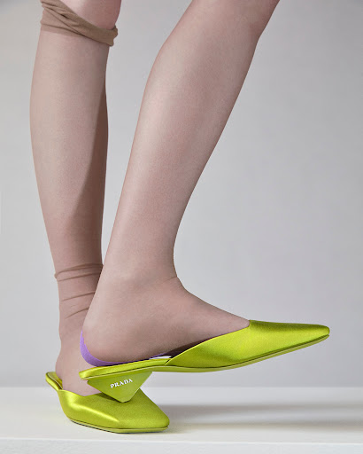 Close-up studio shot of a woman's leg wearing tan-colored hosiery and a chartreuse Prada mule shoe against a neutral background.