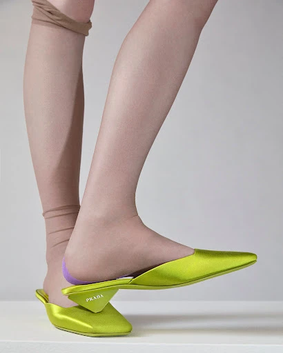 Close-up studio shot of a woman's leg wearing tan-colored hosiery and a chartreuse Prada mule shoe against a neutral background.