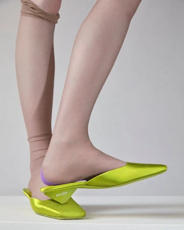Close-up studio shot of a woman's leg wearing tan-colored hosiery and a chartreuse Prada mule shoe against a neutral background.