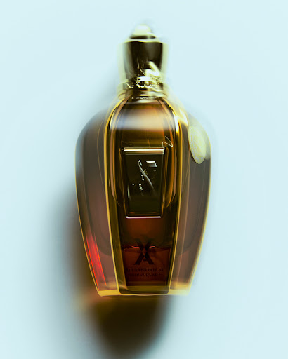 Close-up studio shot of a golden perfume bottle with intricate detailing and a blurred background. The bottle features a clear, ornate design.