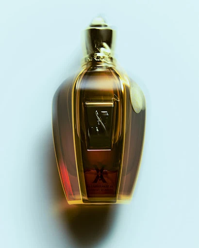 Close-up studio shot of a golden perfume bottle with intricate detailing and a blurred background. The bottle features a clear, ornate design.
