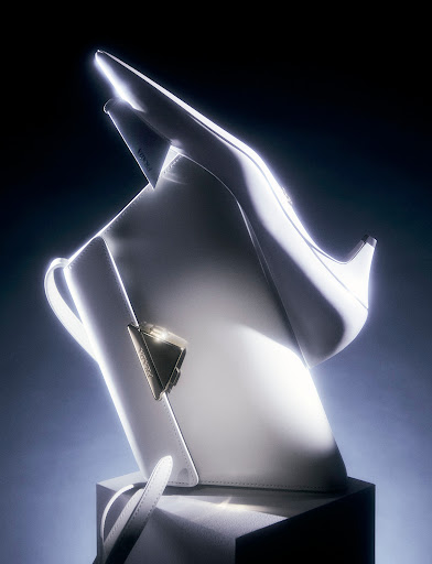 Elevated product shot of a white leather handbag next to a matching stiletto shoe, illuminated by studio lighting against a dark background.