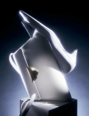Elevated product shot of a white leather handbag next to a matching stiletto shoe, illuminated by studio lighting against a dark background.