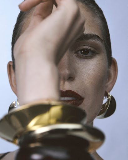 Close-up portrait of a woman with dark lipstick and gold hoop earrings, holding her arm up in the foreground by Marco Gazza.