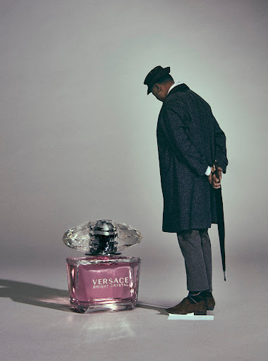 Man in dark coat and hat standing beside a giant Versace Bright Crystal perfume bottle on a neutral studio background