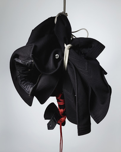 Black fabric garment hung and bound with white and red rope against a neutral light grey studio backdrop.