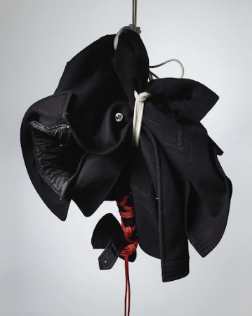 Black fabric garment hung and bound with white and red rope against a neutral light grey studio backdrop.