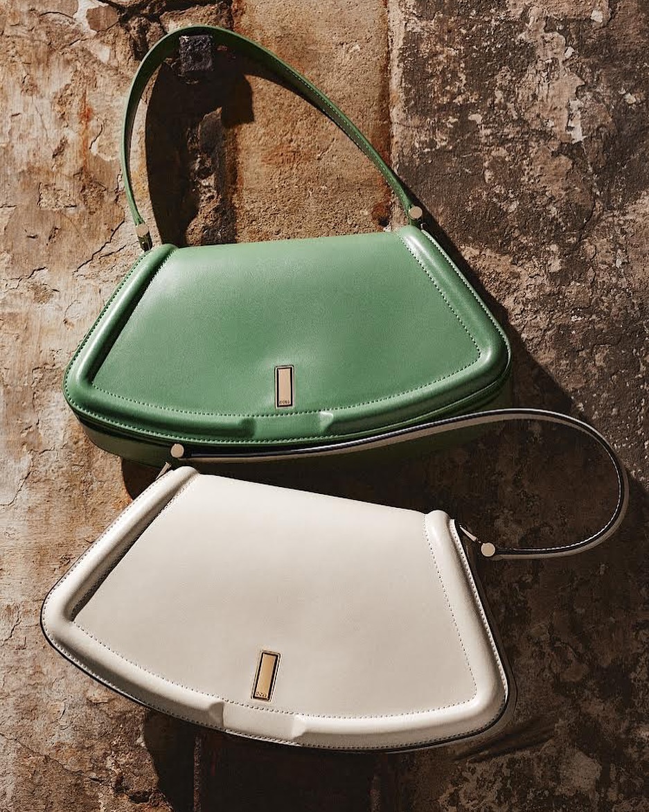 Two stylish leather shoulder bags, one mint green and one off-white, are elegantly displayed against a textured wall backdrop.