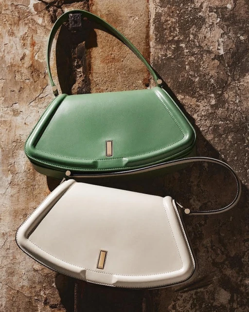 Two stylish leather shoulder bags, one mint green and one off-white, are elegantly displayed against a textured wall backdrop.