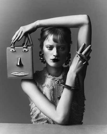 Monochrome portrait of a woman with dark lipstick holding a handbag shaped like a face against a neutral gray background.