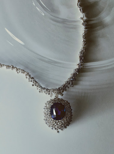Elegant diamond necklace featuring a large oval-cut blue gemstone pendant, set against a softly patterned, light background. Jewellery aesthetic.