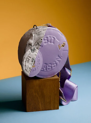 Stylish lavender round purse with strap and decorative coin displayed on a wooden block against an orange and blue background.
