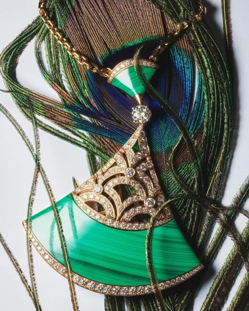Close-up of a green malachite pendant necklace with gold filigree and diamond accents, set against peacock feathers on a white background.