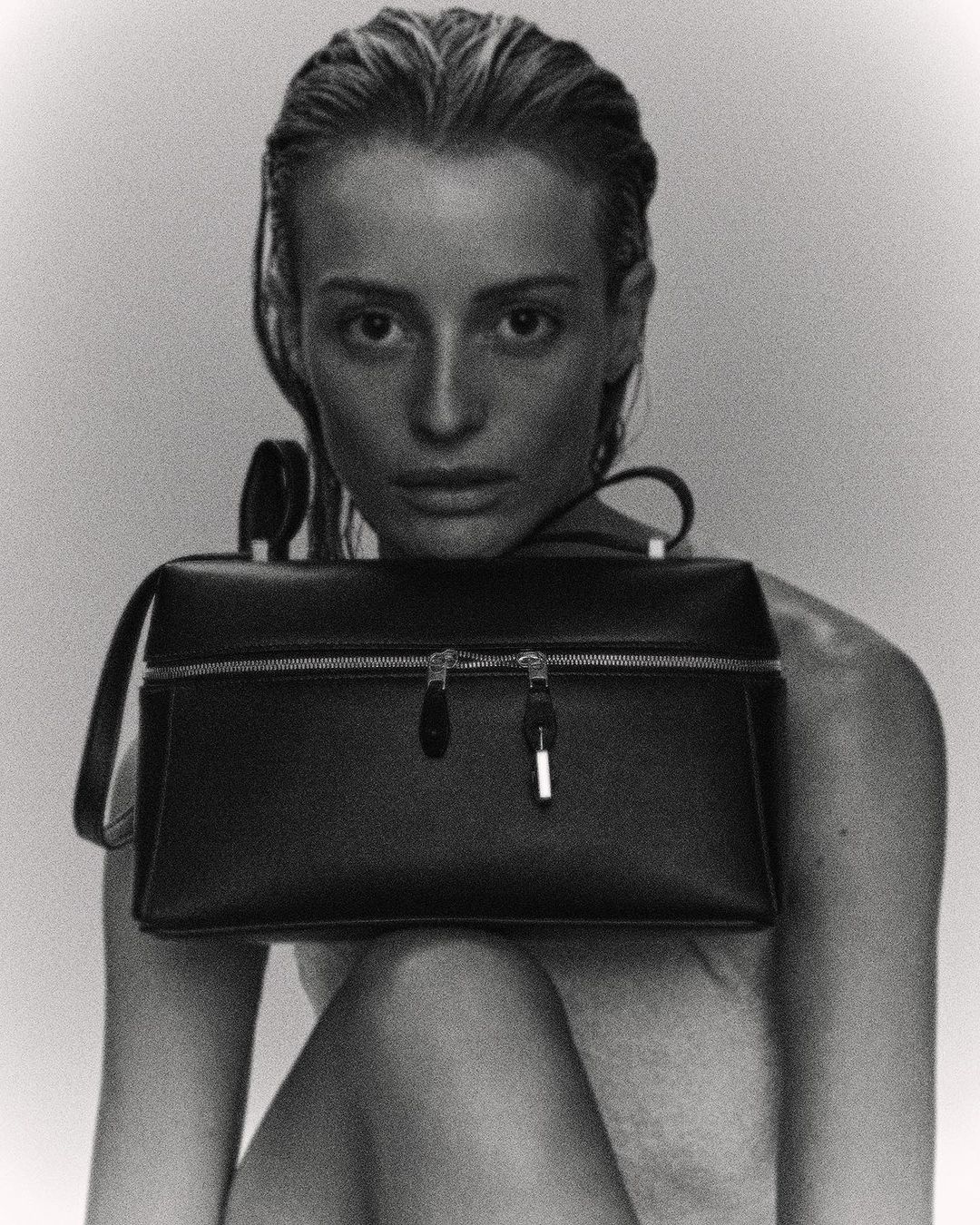 Black and white studio shot of a stylish adult woman holding a structured handbag, with slicked-back hair and soft, diffused lighting.