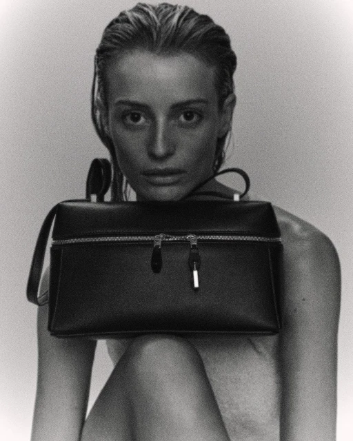 Black and white studio shot of a stylish adult woman holding a structured handbag, with slicked-back hair and soft, diffused lighting.