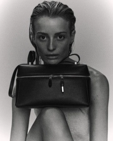 Black and white studio shot of a stylish adult woman holding a structured handbag, with slicked-back hair and soft, diffused lighting.