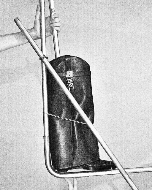 Leather boot suspended on a metal frame with a hand visible in the upper left corner, set against a plain background.