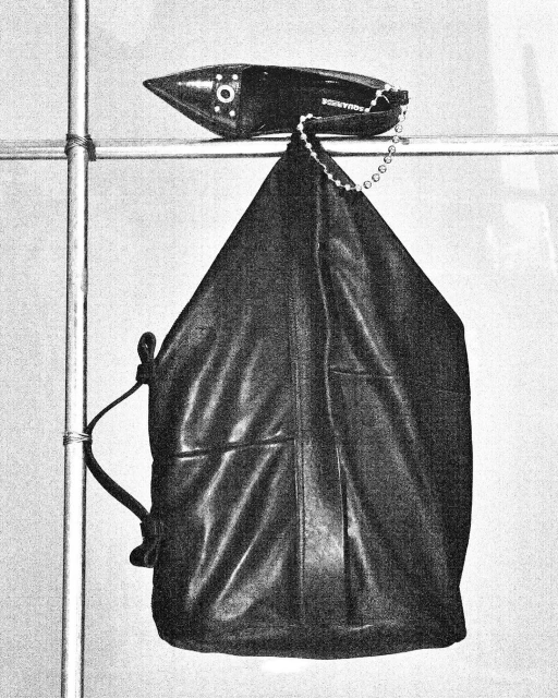 A black leather bag hangs from a metal bar near a black, pointed-toe shoe. The grayscale image shows fashion items in a simple setting.