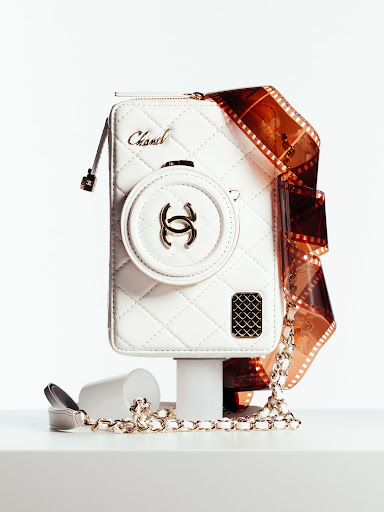 Chanel white leather purse resembling a camera on a white platform with a roll of film and a chain strap. Studio lighting.