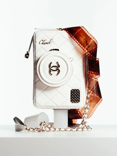 Chanel white leather purse resembling a camera on a white platform with a roll of film and a chain strap. Studio lighting.