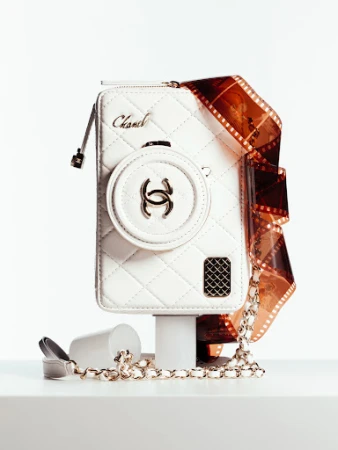 Chanel white leather purse resembling a camera on a white platform with a roll of film and a chain strap. Studio lighting.
