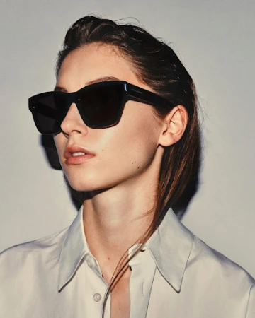 Woman wearing dark oversized sunglasses and a white collared shirt against a neutral grey studio background by Marco Gazza