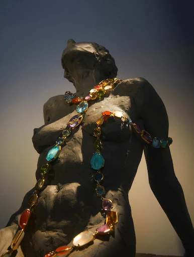 Bust statue of a human form with a colorful gemstone necklace, captured with soft lighting and a low-angle perspective.
