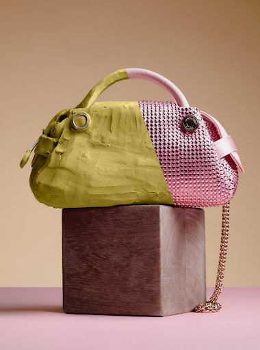 A handbag with one half covered in matte olive clay and the other half in glittering pink rhinestones, resting on a wooden block.