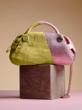 A handbag with one half covered in matte olive clay and the other half in glittering pink rhinestones, resting on a wooden block.