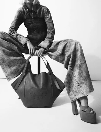 Monochrome fashion portrait of a model in wide-leg acid wash denim holding a large structured leather tote bag against a plain background