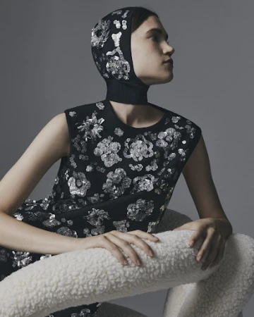 Model wearing a sequined black dress and matching hood, seated behind a white textured chair against a grey backdrop.
