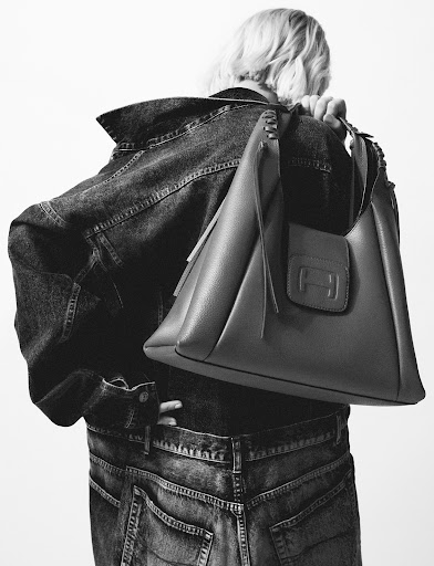 Fashion shot of a woman wearing a denim jacket and jeans carrying a dark leather handbag over her shoulder against a white backdrop.