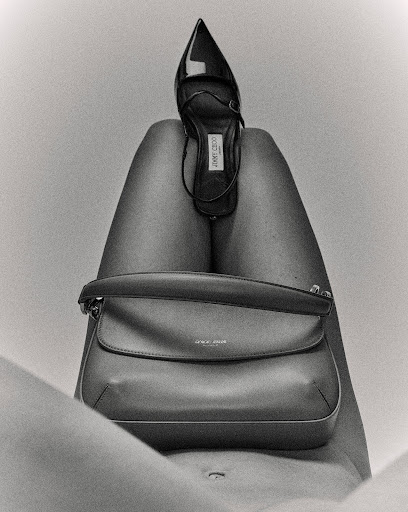 A black and white shot of a woman's legs with a black designer high heel placed on the upper thighs and a handbag on the lower thighs.